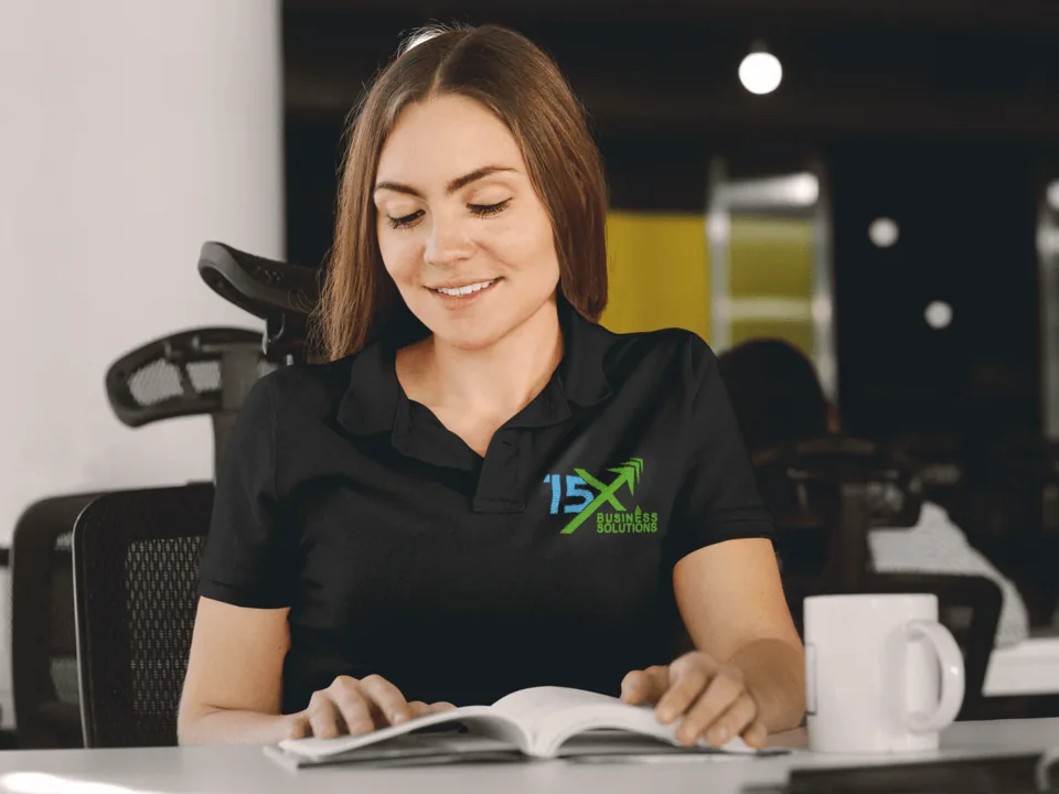 Woman in a black 15X Business Solutions polo shirt reading at an office desk