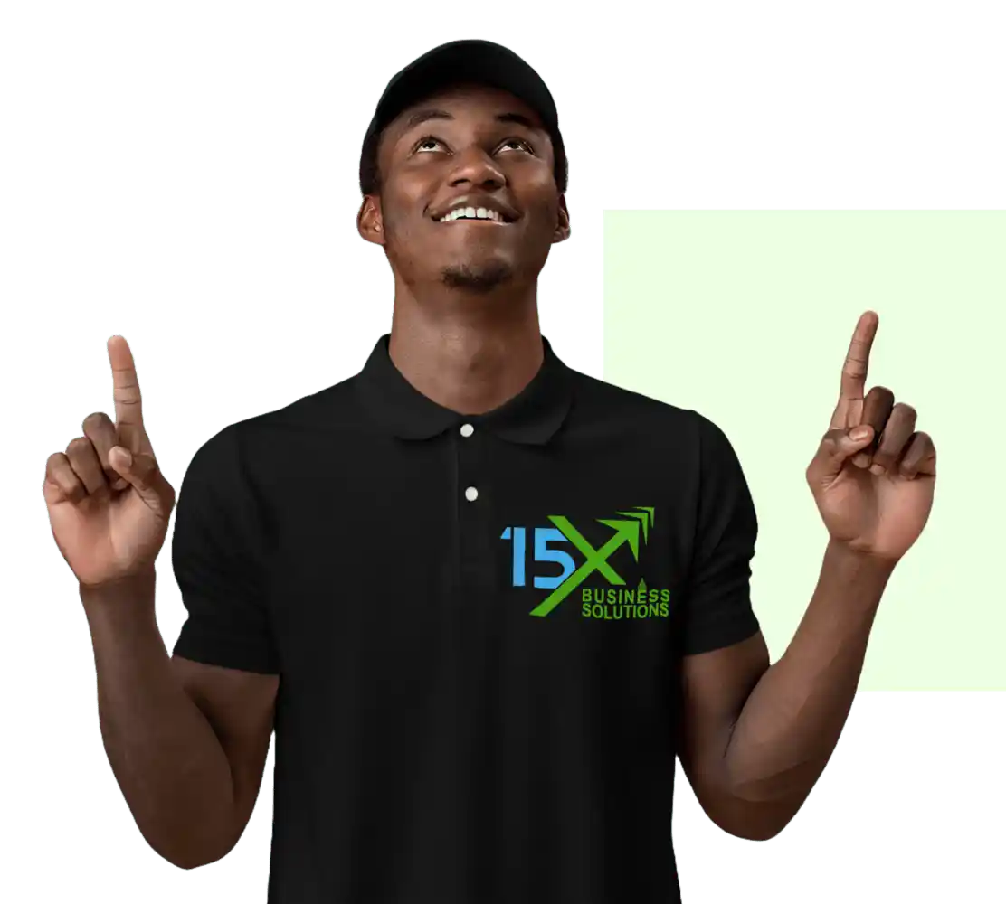 15X Business Solutions team member wearing a black branded polo shirt and cap, smiling and pointing upward with both hands.